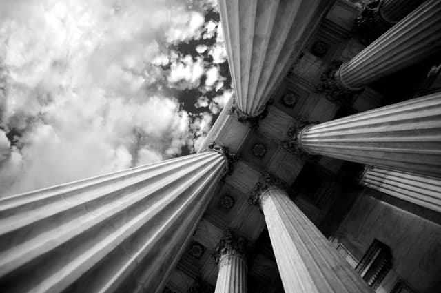 Looking up at tall classical columns supporting a stone portico, with clouds visible above.