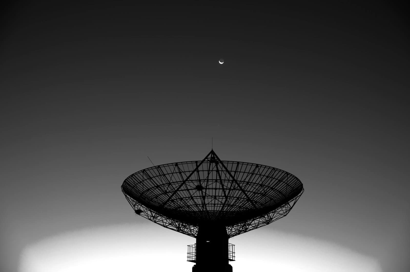 A large satellite dish silhouetted against the sky with a crescent moon above.