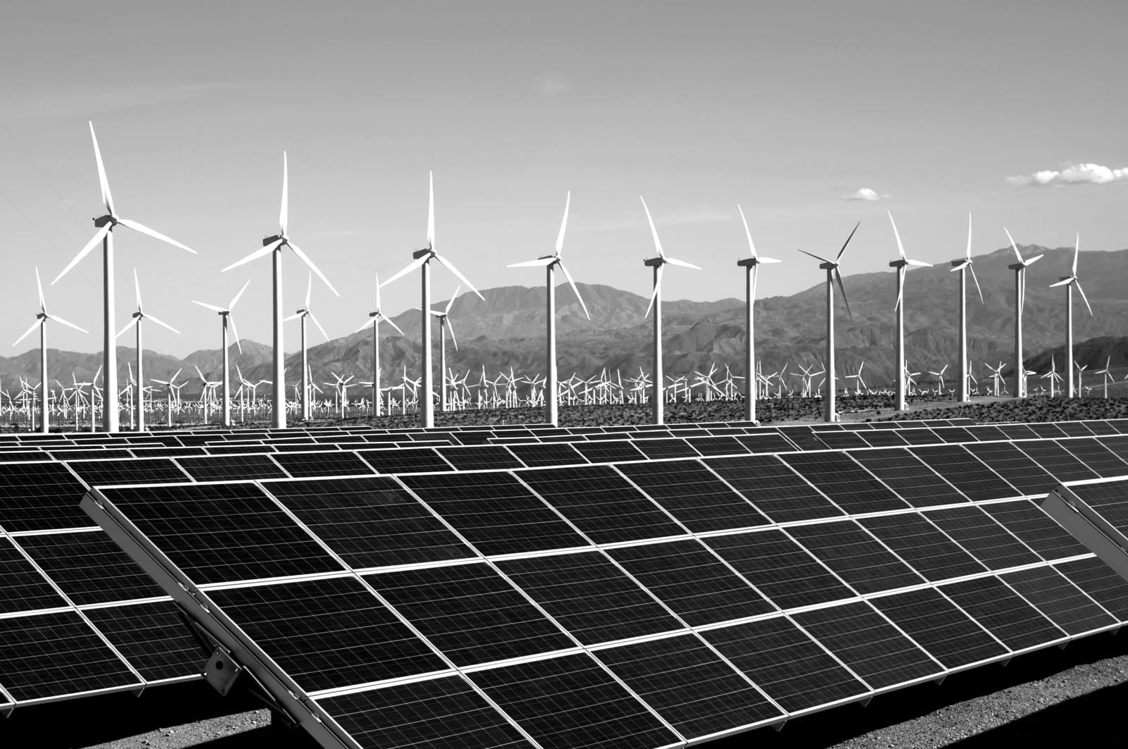Solar panels in the foreground with wind turbines across a renewable energy site, set against a mountainous landscape.