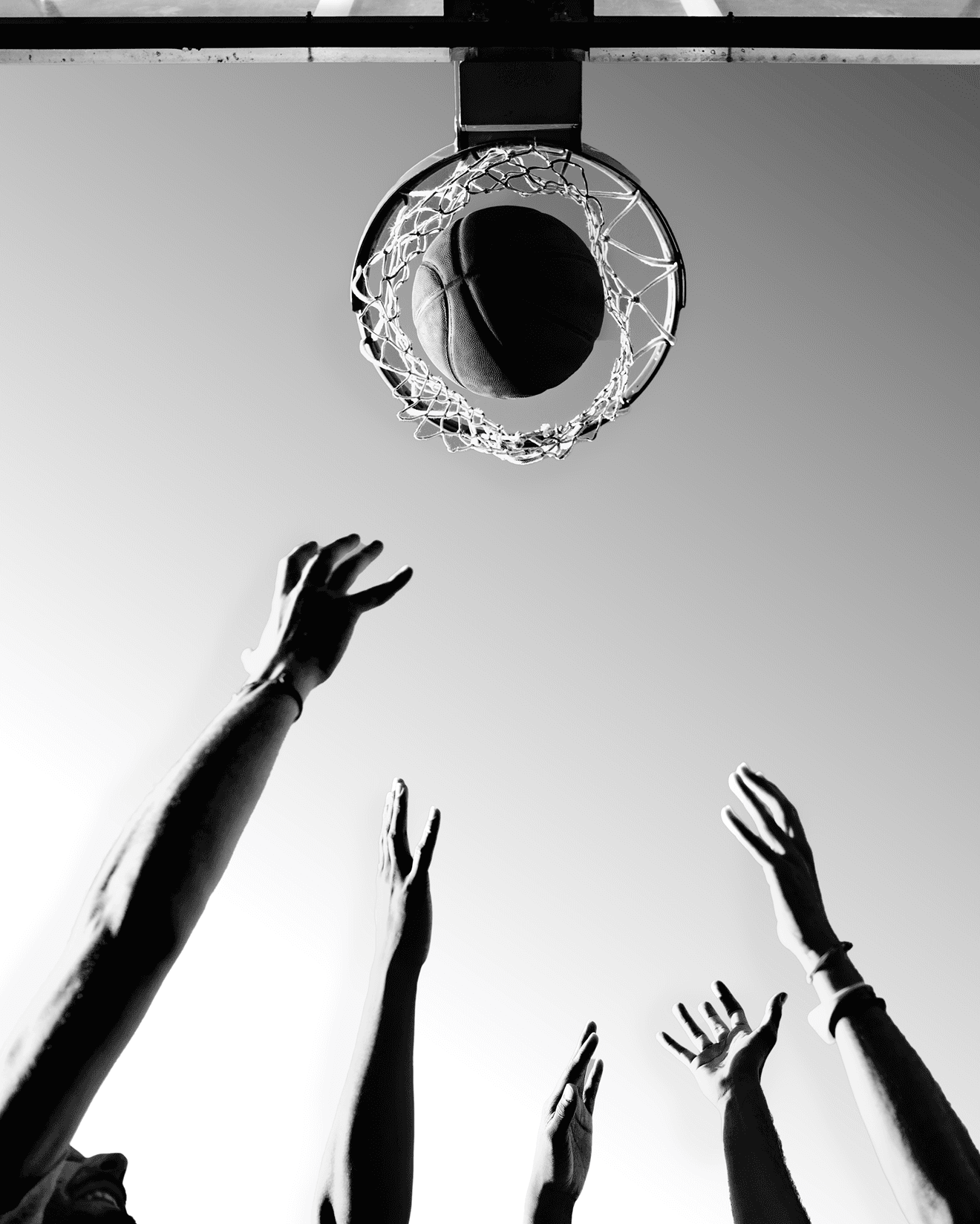 Basketball passing through hoop with multiple hands reaching upward against clear sky, viewed from below.